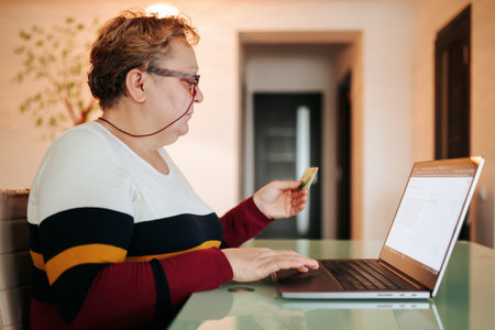 A senior female is shopping online for glasses and other items from the comfort of her home, using her laptop and credit card. She is wearing her eyeglasses and taking advantage e-commerce sites.の写真素材