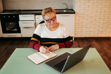 A senior woman sits at home, surrounded by books and papers, as she works on her laptop. She wears glasses as she concentrates on her e-learning course, furthering her education through online study.の写真素材