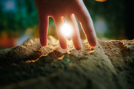 A close-up shot of a human hand digging into the soil, with selective focus on the fingers and the lush garden in the backgroundの写真素材