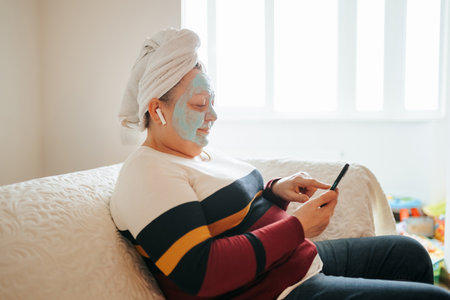 Elderly Woman Enjoying Skincare Routine, A beautiful, overweight senior woman sits in her comfortable living room while wearing a facial mask and listening to music on her smartphone.の写真素材