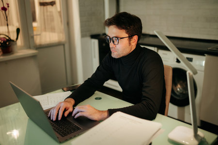 Freelancer at Night Man Working on Notebook Computer in Indoor Setting. Focused male freelancer using his laptop to work at home during the night.の写真素材