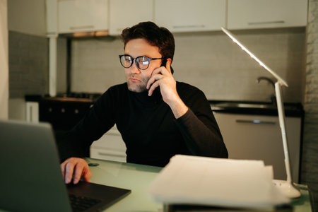 A serious-looking young adult talking on the phone with glasses typing away at his laptop, lost in thought as he works on a project late into the night.の写真素材
