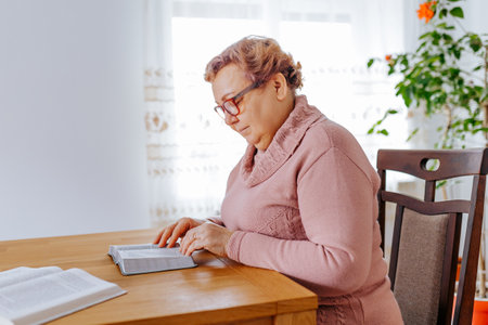 An older woman, glasses on her face, sits in her comfortable armchair at home, lost in the pages of her book. The afternoon sun streams in, casting a warm glow over the scene.の写真素材