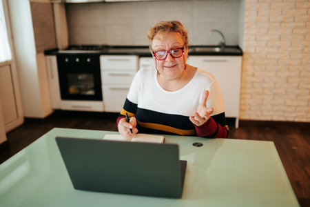 Online Learning for Seniors An aged woman, sits at home with her laptop and eyeglasses. She is engaged in an e-learning session talking, gaining knowledge and information.の写真素材