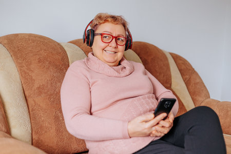 An elderly woman sitting comfortably on her sofa, listening to her favorite tunes with her headphones, looking happy and relaxed at home. Smiling Senior Lady Relaxing on Sofa with Headphones Onの写真素材