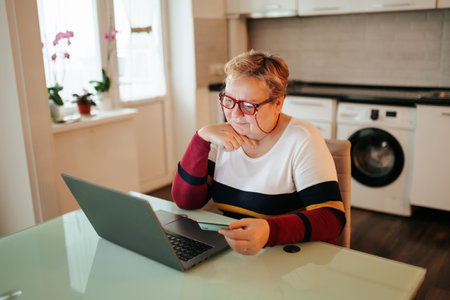 A senior woman, looking overweight, is sitting at home and using her laptop while holding her credit card, making an online purchase through e-commerce.の写真素材