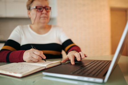 An elderly woman is seen typing away at her laptop while wearing her eyeglasses. She is embracing the convenience of e-education, learning new information and expanding her knowledge from the at homeの写真素材