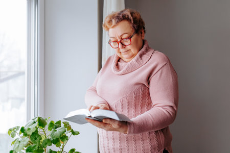 A woman, perhaps overweight or elderly, enjoying the comforts of her home while reading her favorite book next to the window. Her glasses sit atop her head as she intently peruses the pages.の写真素材