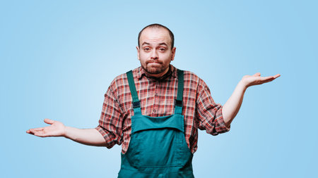 questioning expression on the face of a young man looking at camera, dressed in coveralls and appears to be puzzled, with his hands outstretched in an inquisitive gesture. studio shot blue background.の写真素材