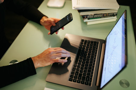 A close-up photo of a salespersons hands using a smartphone to buy and sell stocks on the go, while working from home at night. of hands on a laptop and smartphone for late-night trading at homeの写真素材