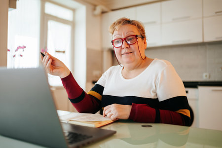 Learning Never Stops A female senior, uses her laptop and glasses to continue her education through e-learning talking. She at home, embracing technology to expand her knowledgeの写真素材