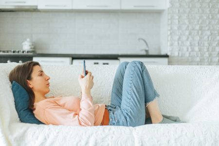 A pretty young woman sits comfortably on her couch at home, absorbed in reading on her smartphone, while enjoying a relaxing evening.の写真素材