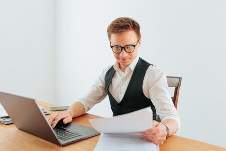 A white-collar man, wearing glasses and a dress shirt, sits at his home office desk, typing on his laptop, with the city skyline visible through the window behind him.の写真素材
