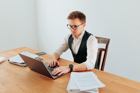 A stylish young businessman, dressed in a white-collar shirt and tie, sits at his modern home office, looking at his laptop screen through his glasses, while taking notes.の写真素材