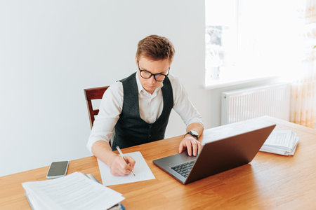 A businessman is working on a project in his modern office, writing down his ideas on a paper while wearing eyeglasses and a white collar shirt. His laptop is open on the desk, and the pen is placedの写真素材