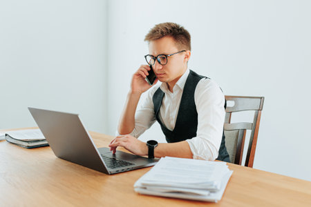 A young Caucasian man in eyeglasses is engaged in a serious conversation on his smartphone while sitting at his white-collar job in the office. In front of him lies a laptop, and behind him,の写真素材