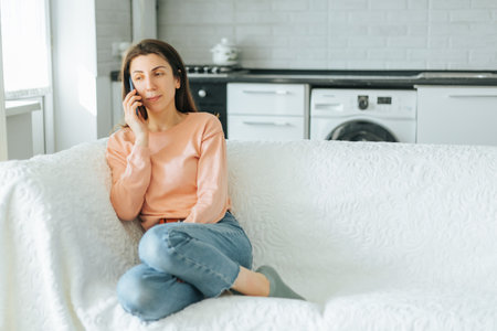 A young woman catching up with her best friend on the phone, sitting on her favorite sofa in her living room. The authentic and laid-back scene is completed by the colorful throw pillowsの写真素材