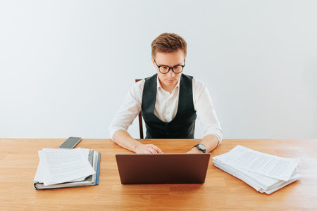 A male office worker in his home office, surrounded by paperwork and his laptop, wears glasses as he concentrates on his work, his white-collar shirt unbuttoned at the neck.の写真素材