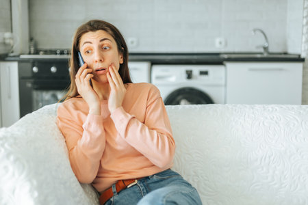 The expression on this young womans face is one of surprise and wonder as she talks on her smartphone, sitting comfortably on her sofa in the living room of her home.の写真素材