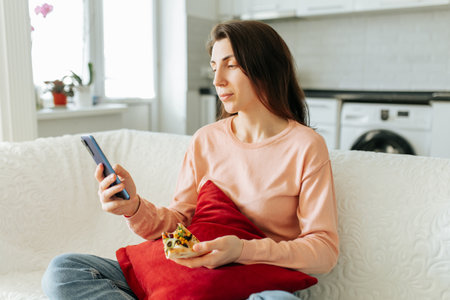 A young woman sits comfortably on her couch, browsing her smartphone with a content expression on her face, while the soft sunlight floods the living room.の写真素材