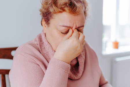Stressed Senior Woman at Home Feeling Sad and Achy While Reading with Glasses. A senior woman sits at home with a book in her hand, her glasses perched on her nose as she struggles to readの写真素材