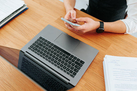 An unrecognizable mans hands typing on a sleek black laptop keyboard while holding a smartphone in his other hand. The close-up shot captures the stylish gadgets on a modern desk in an office.の写真素材