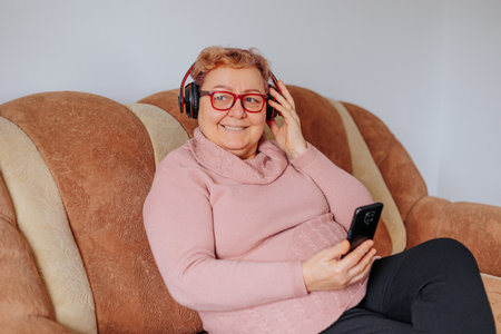 A pretty senior lady relaxing on her sofa at home, listening to her favorite music with headphones and smiling happily. Home Harmony Overweight Senior Lady Smiling with Headphones Onの写真素材