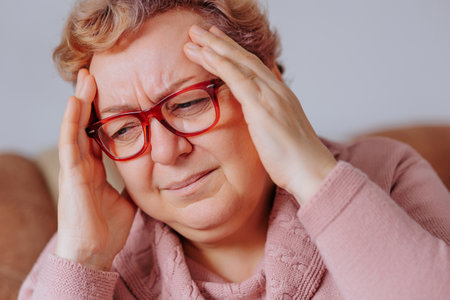Elderly woman struggling with a severe headache, her hands on her head, while sitting in her comfortable living room, Dealing with Difficulty Elderly Woman Battles Aches and Painsの写真素材