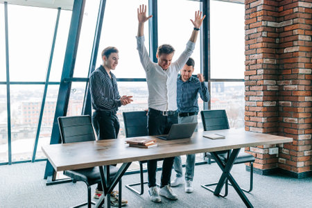 The happiness is palpable in this photo of three successful businessmen, all wearing suits and sharing a moment of delight over a laptop, surrounded by their colleagues and office decor.の写真素材