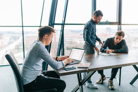 Three young men working on their laptops, with their mentor providing feedback and support in a professional workspace. male mentor providing one-on-one instruction to a trainee on their laptopの写真素材