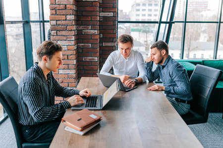 Three guys working together on a team project, all with their laptops open and focused on work in an office environment. team of young professionals discussing business strategies while workingの写真素材
