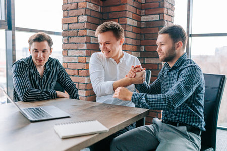 Three young men in a bright office space celebrate their success with high-fives and big smiles, while their laptops and work projects are scattered around them.の写真素材
