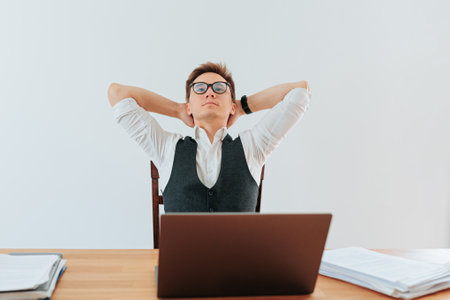 A successful businessman sitting at his desk in his office, looking satisfied and relaxed as he works on his laptop. He is dressed in a white shirt and a navy blue suit, his collar unbuttoned,の写真素材