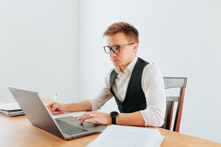 A young businessman, dressed in a white-collar shirt and glasses, works in his home office, typing on his laptop, with a pensive expression on his face.の写真素材