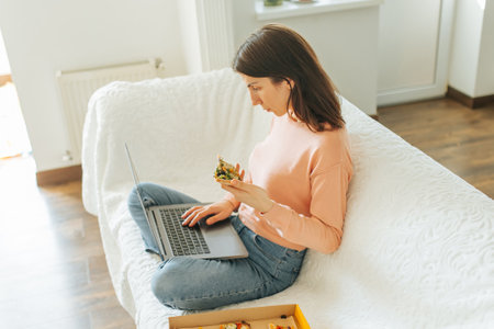 A young woman multitasks by working on her laptop and eating pizza in the comfort of her own home. real and authentic moment, a young woman is captured studying on her laptop while enjoyingの写真素材