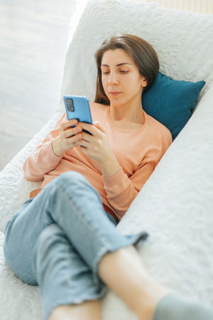 young adult woman is holding her smartphone while sitting on the couch in her living room, browsing through photos and feeling satisfied.の写真素材