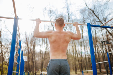 Man Takes on the Challenge of Pull-Ups at Outdoor Gym. In a display of strength, a man does naked pull-ups on a bar outside, showcasing his chiseled physique.の写真素材