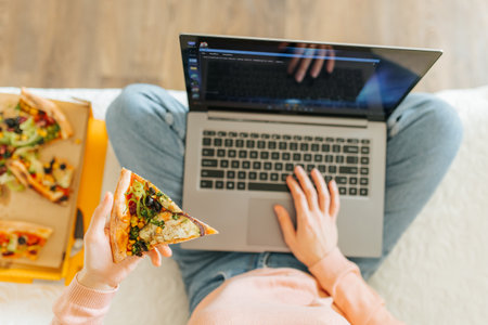 A close-up of a womans hand holding a slice of pizza while she shops for new items online on her laptop. womans hands typing on her laptop while she enjoys a slice of pizza during a break from herの写真素材
