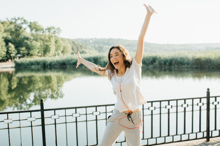 Winner as style of life. Horizontal shot of young beautiful woman in sports clothing smiling while passing finish line during jogging. Evening sunlight on background, Sport lifestyle.の写真素材