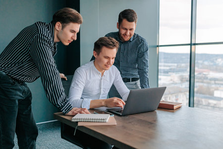 Three young men sitting in an office, working on their laptops and collaborating on a project. group of people in business attire, including a businessman and his two colleagues, all typing awayの写真素材