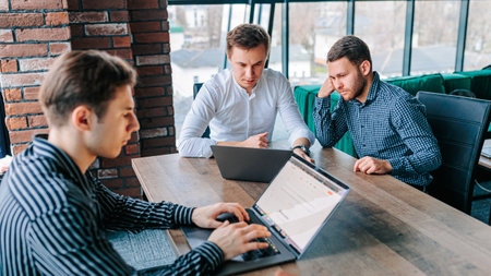 A male mentor offering guidance to a trainee who is working on a laptop in an office environment. Three guys busy working on their laptops, while their trainer stands behind them to supervise.の写真素材