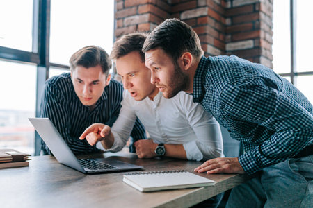 Three young men in an office, staring in amazement at their laptop screens. The work in front of them seems to have left them flabbergasted and bewildered.の写真素材