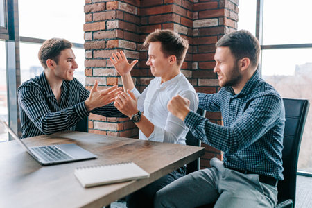 In this top-view photo of a group of three men in a modern office, they are all sitting around a laptop with delighted expressions, celebrating the success of their latest project.の写真素材