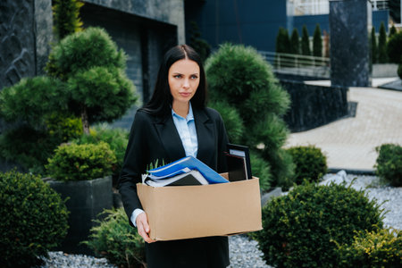 A person holding a box, outside their former workplace, after being fired and feeling jobless. Starting Over Woman Outside with Box of Possessions After Job Lossの写真素材