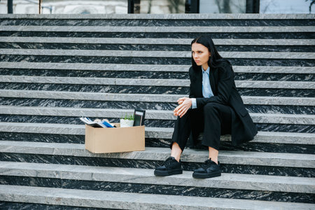 A frustrated woman sits on the stairs outside, feeling the weight of her jobless situation and the struggle to make ends meet. Depressed Woman with Cardboard Box on Stairsの写真素材