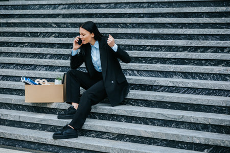 An upset unemployed woman speaking on her mobile phone while sitting on the stairs. Emotional Phone Call Speaking About Unemployment and Loss of Workの写真素材