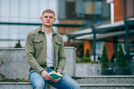 This portrait of a young male with a book captures his intense concentration as he sits in an urban setting. City Life and Literature A Handsome Man Sitting with Book Outsideの写真素材