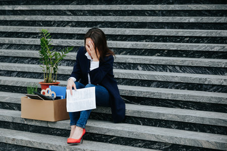 depicts a middle-aged woman sitting on a stair, her face etched with worry and unhappiness, symbolizing the personal struggle caused by job loss, unemployment, and the loss of work.の写真素材