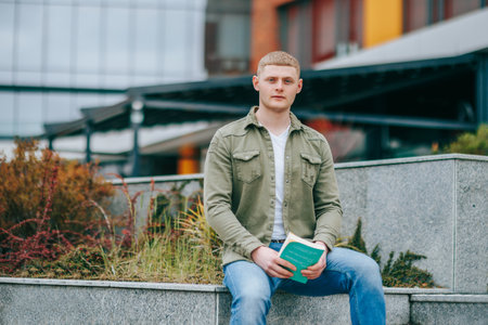 This young male is engrossed in his book while sitting in an urban location, the portrait capturing his intense concentration. Book Lovers Retreat A Handsome Man Sitting Outdoors with Bookの写真素材