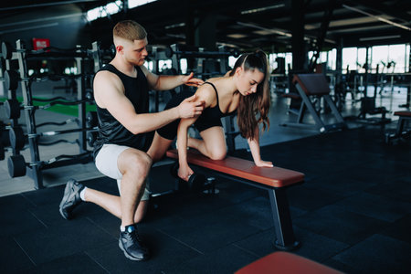 In the gym, a skilled male personal trainer aids a woman in her weightlifting journey, specifically focusing on her dumbbell lift and technique.の写真素材